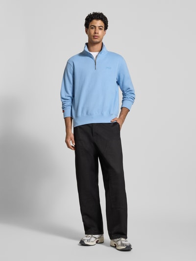 Männermodel trägt Regular Fit sweatshirt mit logostitching van Superdry in lichtblauw, katoenmix, opstaande kraag met ritssluiting, elastische boorden, ruglengte maat M 65 cm, mouwlengte 63 cm, ganzkörperansicht van voren.