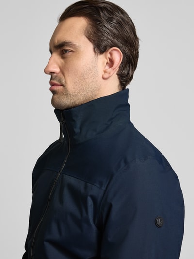 Męski model nosi Regular Fit Kurtka ze stójką Model 'Alvin' od Didriksons, zamek błyskawiczny, elastyczne wykończenia, wpuszczane kieszenie, kolor granatowy, tekstura gładka, długość tyłu w rozmiarze M: 69 cm, szerokość ramion w rozmiarze M: 44 cm, szerokość klatki piersiowej w rozmiarze M: 54 cm, długość rękawów w rozmiarze M: 70 cm, detaliczna bliska perspektywa lewy profil.