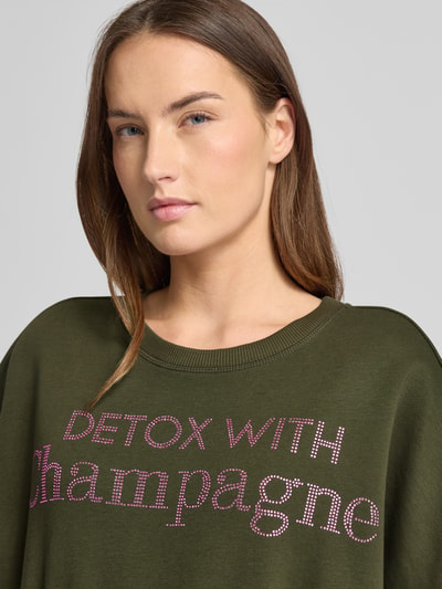 Kobiece model prezentuje Oversized Sweatshirt aus reiner Baumwolle od miss goodlife, kolor ciemnozielony, nadruk z napisem, okrągły dekolt, krój oversized, wykończenia elastyczne, bawełna, detal: nadruk z napisem, ujęcie: zbliżenie górnej części ciała.