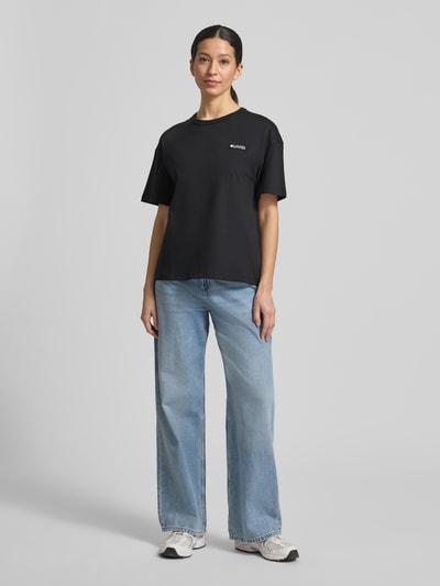 Frauenmodel trägt Damen T-Shirt aus reiner Baumwolle mit Modell 'CSC™' von Columbia in Schwarz mit Label-Print, lockerer Passform, Kurzärmelig, Rundhalsausschnitt, Omni-Shade Technologie UPF 30, Rückenlänge bei Größe S 60 cm, Ganzkörperansicht von vorne.