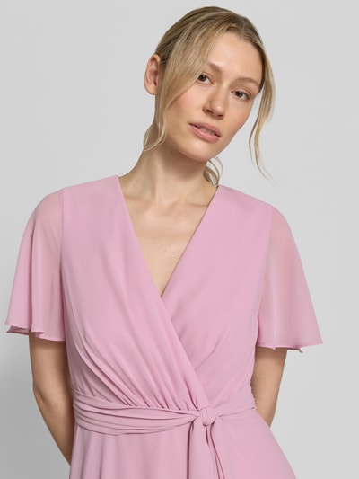 Vrouwenmodel draagt knielanges Cocktailkleid in Wickel-Optik van Christian Berg Cocktail, lichtroze chiffon, a-lijn, V-hals, bindriem, knielengte, borstwijdte 33 cm, ruglengte 100 cm, mouwlengte 22 cm. Close-up bovenlichaam.