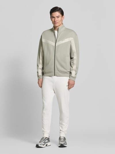 Męski model nosi Relaxed Fit Sweatjacke mit Baumwoll-Anteil Modell 'DARTI' od HUGO, jednolity kolor, długi rękaw, zamek, ściągacze, boczne kieszenie, detal z logo, taśma chevron, stójka; widok całej sylwetki z przodu.