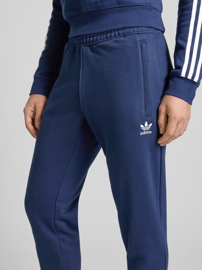 Männermodel trägt 'Sweatpants mit elastischem Bund' von adidas Originals in dunkelblau, lockere pasvorm, katoenmix, label-stitching, Franse steekzakken, elastische tailleband, detailopname heup.