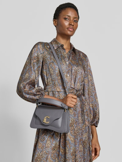 Frauenmodel trägt Shoulder Bag mit Strukturmuster von Coccinelle in Grau mit Logo-Detail und verstellbarem Schulterriemen, Ganzkörperansicht von vorne. Geteiltes Hauptfach mit Überschlag, Maße 23x18x10 cm, Länge Schulterriemen 38 cm.