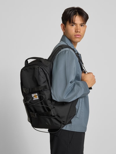 Männermodel trägt Rucksack mit Logo-Patch Modell 'KICKFLIP' von Carhartt Work In Progress in Schwarz, Reißverschlusstasche, Nahaufnahme Oberkörper.
