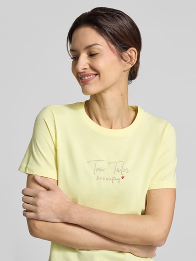 Modelka kobieca nosi Regular Fit T-Shirt aus Bio-Baumwoll-Mix od Tom Tailor, kolor jednolity, krótki rękaw, okrągły dekolt, napis z logo, miękka tekstura, ujęcie z przodu zbliżenie na górną część ciała.