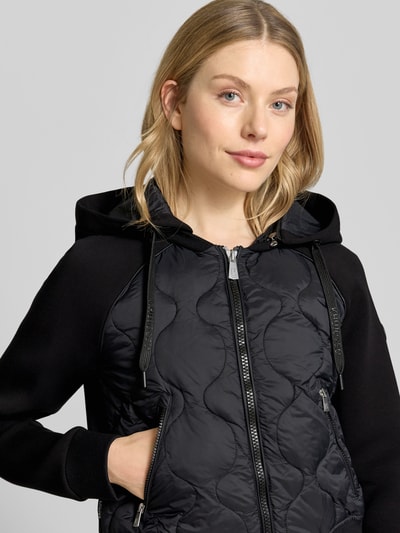 Frauenmodel trägt Steppjacke mit Reißverschlusstaschen Modell 'MALAYA' von khujo in Schwarz, Kombination aus glatter und gesteppt strukturierter Oberfläche, Regular Fit, Kapuze, Nahaufnahme Oberkörper. Rückenlänge, Schulterbreite, Brustweite und Ärmellänge entsprechend Größe S.