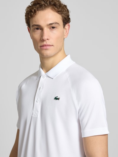Męski model nosi Regular Fit Sport Poloshirt mit Label Patch od Lacoste Sport, biała piqué, jednolity kolor, krótki rękaw, kołnierzyk polo, zapięcie na trzy guziki, aplikacja z logo, całkowite ujęcie górnej części ciała z przodu.