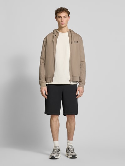 Męski model nosi Slim Fit Sweatjacke mit Kapuze od EA7 Emporio Armani w beżu, krój slim fit, przód, widok całej sylwetki.