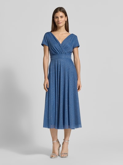 Modelka kobieca nosi Schimmerndes Cocktailkleid mit V-Ausschnitt od Vera Mont, tiul, linia A, sukienka midi, efekt błyszczący, dekolt w serek, długość tyłu w rozmiarze 36: 100 cm, szerokość w rozmiarze 36: 42 cm, widok całej sylwetki z przodu.