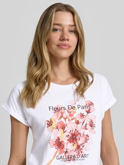 Frauenmodel trägt Damen T-Shirt mit Motiv-Print Modell 'RINA' von Soyaconcept aus Baumwoll-Elasthan-Mix, Rundhalsausschnitt, Regular-Fit, weiß mit floralem Print, Ärmel mit Aufschlag, Rückenlänge bei Größe S: 61 cm, Nahaufnahme Oberkörper.