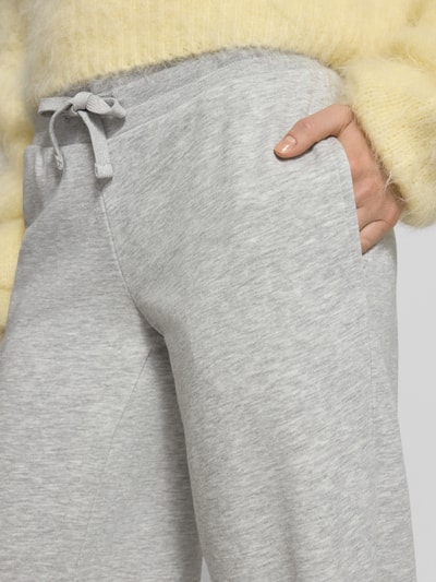 Kobiece modelka nosi Wide Fit Sweatpants mit Viskose-Anteil od REVIEW, mieszanka bawełny i wiskozy, krój wide fit, elastyczny pas, tunel, długość wewnętrzna nogawki w rozmiarze S: 89 cm, jasnoszary melanż, miękka tekstura, zbliżenie na biodra i rękę w kieszeni