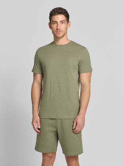 Männermodel trägt Regular Fit T-Shirt aus reiner Baumwolle von Lacoste in Olivgrün, unifarben, Label-Tape, Rundhals, Kurzarm, Ganzkörperansicht von vorne.