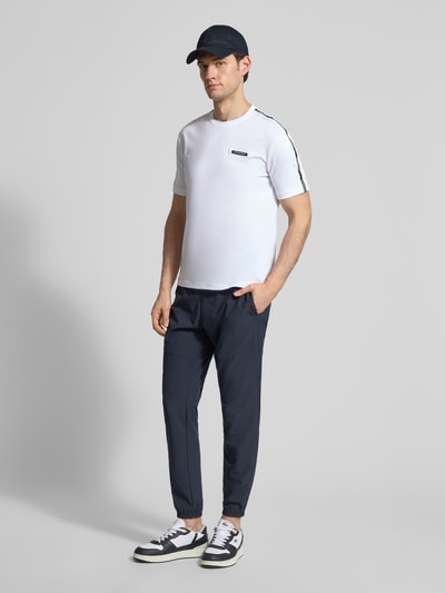 Męski model nosi Regular Fit T-Shirt mit Logo-Patch od EA7 Emporio Armani, biała bawełna, krój regular fit, widok całej sylwetki z przodu.