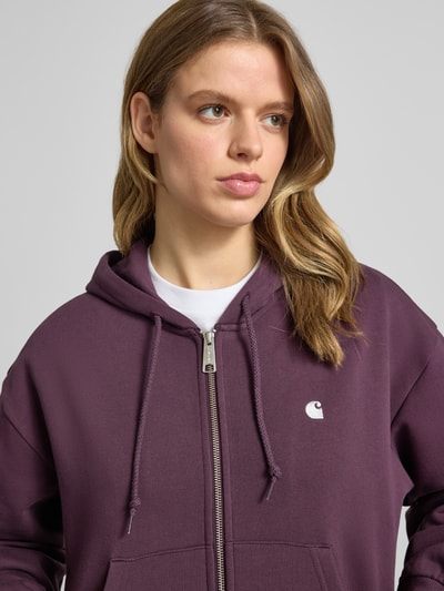 Kobiece model nosi bluzę rozpinaną z wyhaftowanym logo model 'Casey' od Carhartt Work In Progress z bawełny, krój loose fit, kaptur, zamek błyskawiczny, wpuszczane kieszenie, elastyczne wykończenia, widok z przodu w zbliżeniu na górną część ciała.