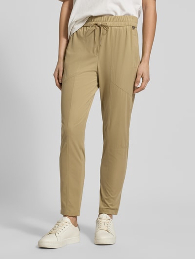 Kobieta nosi Slim Fit Stoffhose mit Reißverschlusstaschen von Marc Cain, beżowy kolor, elastyczny materiał, krój slim fit, długość nogawki 68 cm, szerokość w rozmiarze 36: 37 cm, gumka i tunel, widoczne detale, zdjęcie całej sylwetki z przodu.