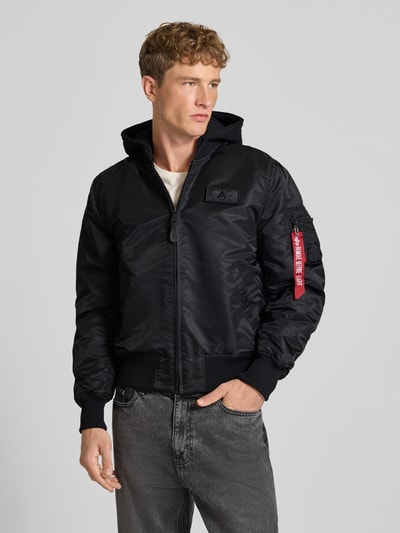 Männermodel trägt Regular Fit Bomberjacke mit Kapuze von Alpha Industries in Schwarz, Webstoff mit Label-Patch, Ganzkörperansicht von vorne.