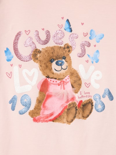 Mädchen trägt Dziecięcy t-shirt z mieszanki bawełny i elastanu Guess z detalem z logo, okrągły dekolt, krój regular fit, różowy, długość tyłu 44 cm przy rozmiarze 116, nadruk z misiem i motylami, Ganzkörperansicht von vorne.
