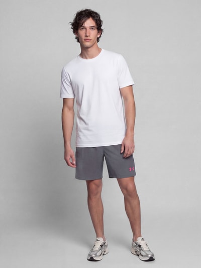 Männermodel trägt Regular Fit Sportshorts mit Logo-Print von Under Armour in Grau mit rosa Logo, kombiniert mit weißem Shirt und Sneakers, Ganzkörperansicht von vorne. Herren Sportshorts von Under Armour.