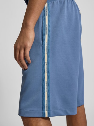 Männermodel trägt Sweatshorts mit Label-Details und Eingrifftaschen von EA7 Emporio Armani, Baumwoll-Mix, Nahaufnahme Oberschenkel. Relaxed-Fit, elastischer Bund, Bundweite 47 cm, Innenbeinlänge 29 cm, Farbe Blau, strukturierte Oberfläche.