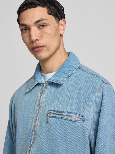 Mężczyzna model nosi Regular Fit Jeansjacke aus reiner Baumwolle Modell 'ELZO' von HUGO Blue, jasnoniebieski denim, tekstura tkaniny, zamek błyskawiczny, label patch, kieszeń na piersi, długi rękaw z mankietem, krój regular fit, zbliżenie na górę sylwetki