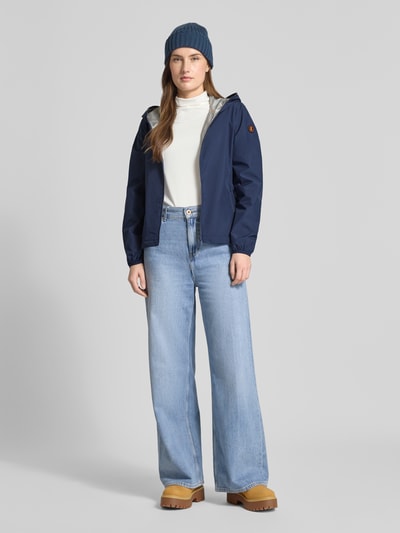 Frauenmodel trägt Regular Fit Jacke mit Zweiwegereißverschluss Modell 'Stella' von SAVE THE DUCK in Dunkelblau, kombiniert mit heller Jeans und Boots, Durchgehender Zweiwegereißverschluss, Eingrifftaschen, Rückenlänge 60 cm, Schulterbreite 42 cm, Brustweite 52 cm, Ärmellänge 64 cm, Ganzkörperansicht von vorne.