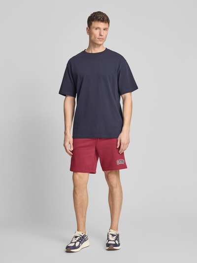 Männermodel trägt Sweatshorts mit Logo und elastischem Bund von GAP in Rot, aus Baumwoll-Mix mit Regular-Fit, Eingrifftaschen, Ganzkörperansicht von vorne.