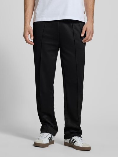 Männermodel trägt Trackpants mit Längsnähten Modell 'Motus' von Pegador, czarne spodnie, materiał z bawełną, krój relaxed fit, elastyczny pas, widoczna tekstura i szwy, zdjęcie prezentuje dolną część ciała, Ganzkörperansicht von vorne.