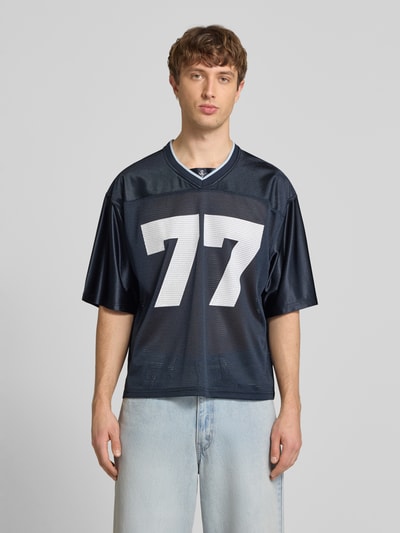 Männermodel trägt T-Shirt in semitransparenter Optik von REVIEW, dunkelblau mit weißer Zahl, V-Ausschnitt, Halbarm, Ganzkörperansicht von vorne.