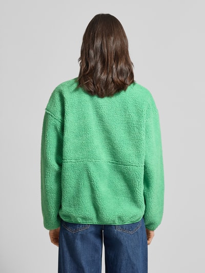 Frauenmodel trägt Jacke aus Teddyfell Modell 'HOKTOWN' von American Vintage, leuchtendes Grün, lockerer Schnitt, Rückenansicht, Nahaufnahme Oberkörper, Teddyfell-Struktur, Reißverschluss, Label-Detail, Eingrifftaschen, Rückenlänge 65 cm, Schulterbreite 60 cm, Brustweite 59 cm, Ärmellänge 55 cm.