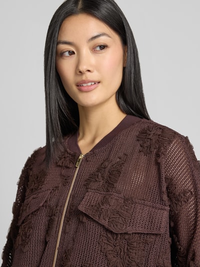 Vrouwenmodel draagt blouson met borstzakken en applicaties van zero, materiaalmix met stretch, loose fit, opstaande kraag, ritssluiting, ruglengte bij maat 36: 54 cm, borstwijdte bij maat 36: 64 cm, mouwlengte bij maat 36: 47 cm, bruine tint, tekstuur met bloemenapplicaties, netstructuur, nahaufnahme bovenlichaam.
