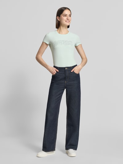 Frauenmodel trägt Slim Fit T-Shirt mit Strasssteinbesatz von Guess aus elastischem Baumwoll-Mix, Rundhalsausschnitt, Logo-Detail, Rückenlänge 59 cm, Ganzkörperansicht von vorne.