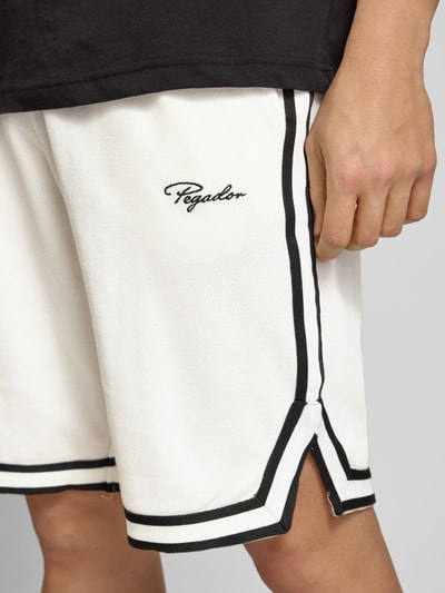 Mężczyzna nosi Shorts mit Logo-Stitching Modell 'Signar' von Pegador z czystej bawełny, biały z czarnymi paskami i logo, elastyczny pas, szerokość w rozmiarze M 40 cm, długość wewnętrzna 27 cm, krój prosty, zbliżenie na górną część nogi