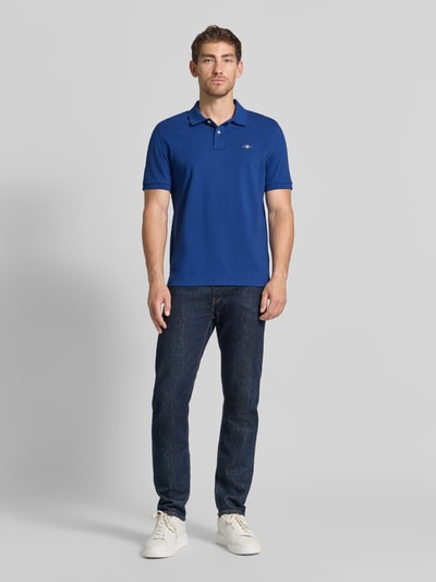 Männermodel trägt Regular Fit Poloshirt aus reiner Baumwolle von GANT in kräftigem Blau, strukturierte Oberfläche, Ganzkörperansicht von vorne.