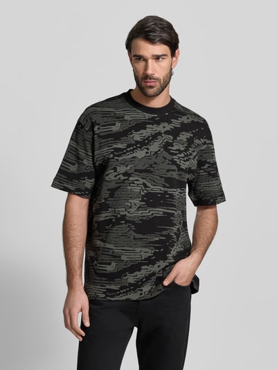 Männermodel trägt Regular Fit T-Shirt aus reiner Baumwolle Modell 'TE_WILDNATURE' von BOSS Orange mit abstraktem Allover-Muster in Schwarz und Oliv, kurzarm, Rundhals, Feinstrick; Ganzkörperansicht von vorne.