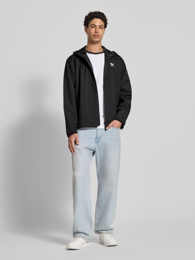 Męski model nosi Kurtka mit Kapuze von adidas Originals, czarna, krój regular fit, wpuszczane kieszenie, nadruk z logo, długość tyłu 71 cm, szerokość ramion 55 cm, szerokość w rozmiarze M 61 cm, długość rękawów 66 cm, zamek błyskawiczny, kaptur, cała sylwetka z przodu