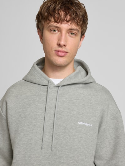 Męski model nosi Hoodie mit Kapuze od Carhartt Work In Progress, szary, luźny krój, bawełna, kaptur, kieszeń kangurka, haft z logo, długość tyłu 70 cm, długość rękawa 62 cm, widok z przodu, zbliżenie na górną część ciała.