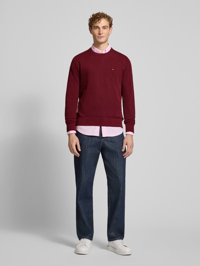 Męski model nosi Regular Fit Pullover aus reiner Baumwolle von Tommy Hilfiger w kolorze bordowym, z fakturą dzianiny, całkowity widok z przodu.