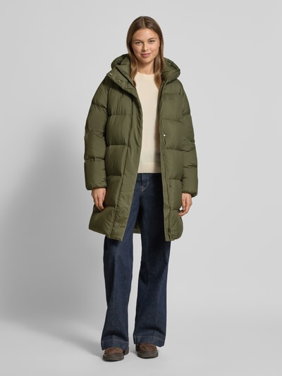 Modelka kobieca ma na sobie Regular Fit Daunenmantel mit Kapuze od Tommy Hilfiger, oliwkowy kolor, długie rękawy, krój regular fit, stojący kołnierz z kapturem, wodoodporna tkanina, boczne kieszenie na zatrzaski, logo na rękawie, widok całej sylwetki z przodu.