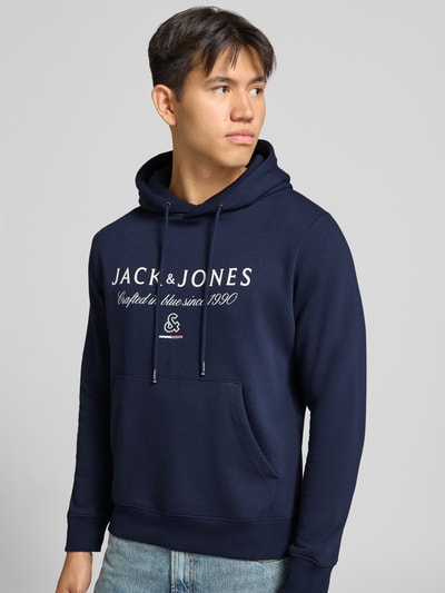 Männermodel trägt dunkelblauen Relaxed Fit Hoodie aus Baumwoll-Mix Modell 'KYLE' von JACK & JONES, Schriftzug vorn, Nahaufnahme Oberkörper. Herren Hoodie, Kapuze mit Tunnelzug, Label-Detail, Känguru-Tasche, Rückenlänge 67 cm, Ärmellänge 65 cm.