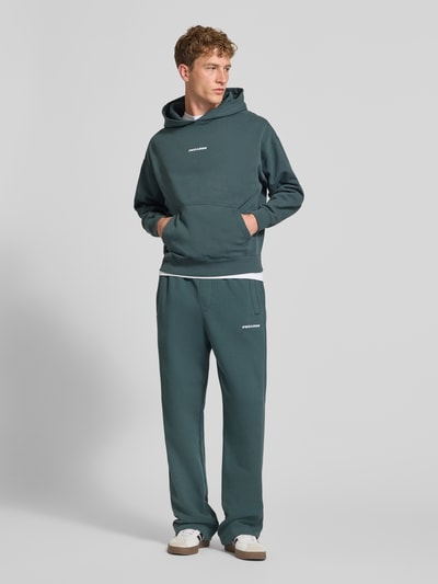 Männermodel trägt Loose Fit Sweatpants aus Baumwoll-Mix mit Label-Detail von PEGADOR in Dunkelgrün, elastischer Bund, Eingrifftaschen, Ganzkörperansicht von vorne.
