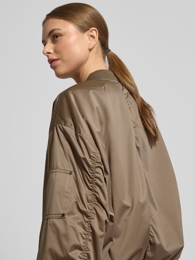 Frauenmodel trägt Regular Fit Bomberjacke mit Stehkragen Modell 'Lorraine' von Guess in Braun mit elastischen Abschlüssen und Reißverschluss, Nahaufnahme Oberkörper. Damen Bomberjacke mit Eingrifftaschen, Rückenlänge 56 cm, Schulterbreite 58 cm, Brustweite 65 cm, Ärmellänge 57 cm.
