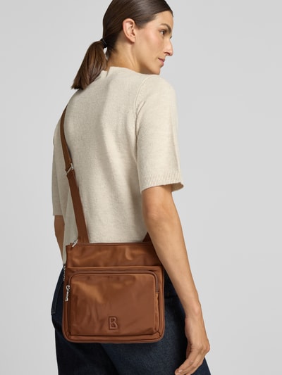 Frauenmodel trägt Handtasche mit Logo-Detail Modell 'Serena' von BOGNER in Braun, glatte Struktur, Sicht auf Hauptfach und Schulterriemen, Ganzkörperansicht Rückansicht. Hauptfach mit Reißverschluss, verstellbarer Schulterriemen, Maße 25x24x2 cm, Länge Schulterriemen 118 cm.