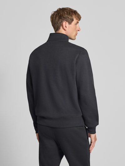 Męski model nosi Sweatanzug mit Label-Details od Champion, mieszanka bawełny, prosty krój, stójka, elastyczny pas, wpuszczane kieszenie, czarny kolor, widoczne logo, zdjęcie całej sylwetki z tyłu, Szerokość w rozmiarze M: 76,42 cm, Długość tyłu w rozmiarze M: 70 cm, Szerokość ramion w rozmiarze M: 51 cm, Długość rękawów w rozmiarze M: 69 cm, Długość wewnętrzna nogawki w rozmiarze M: 76 cm. Ganzkörperansicht Rückansicht