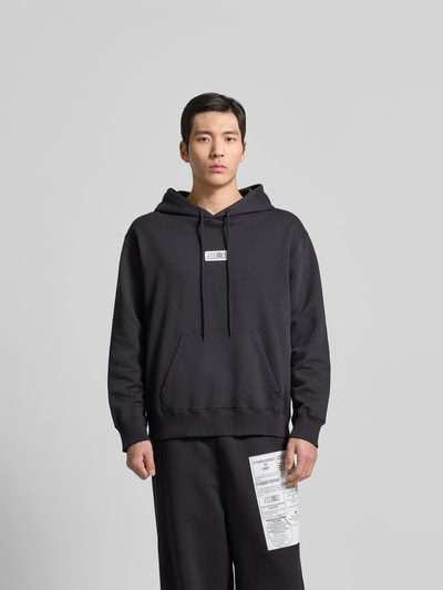Männermodel trägt Hoodie mit Label-Stitching von MM6 Maison Margiela, schwarze Baumwolle, Känguru-Tasche, lockere Passform, Ganzkörperansicht von vorne.