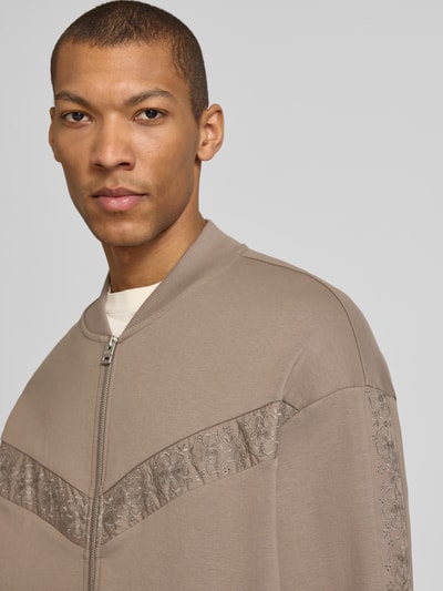 Männermodel trägt Oversized Sweatjacke aus Baumwoll-Mix Modell 'DROMZIP' von HUGO in Taupe mit Logo-Patch und Motiv-Stitching, Nahaufnahme Oberkörper.