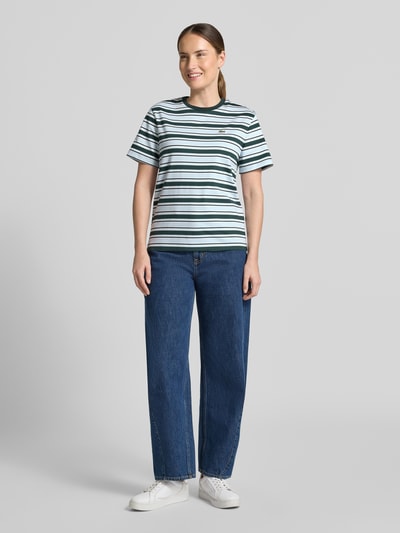 Frauenmodel trägt Regular Fit T-Shirt aus reiner Baumwolle von Lacoste in Weiß mit grünen und schwarzen Streifen, kombiniert mit blauer Jeans, Ganzkörperansicht von vorne. Damen T-Shirt von Lacoste.