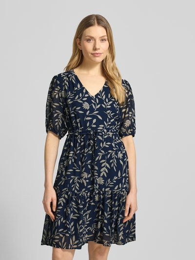 Kobiece model prezentuje Knielanges Kleid mit Kordelzug an der Taille od Apricot, granatowa sukienka z jasnym wzorem liści, dekolt w serek, rękaw o dł. 1/2 z elastycznym zakończeniem, długość do kolan, talia z trokiem, dół obszyty falbaną, fason taliowany, Szerokość w rozmiarze S: 43 cm, długość tyłu w rozmiarze S: 93 cm, długość rękawów w rozmiarze S: 30 cm, zdjęcie całego przodu modelki.