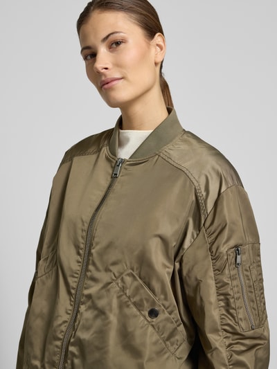 Vrouwenmodel draagt dames boxy fit jack met opstaande kraag, tweewegritssluiting, steekzakken met drukknoopsluiting, model 'JODY' van Beaumont Amsterdam in olijfkleur met gladde textuur, ruglengte maat 36: 54 cm, schouderbreedte: 42 cm, borstwijdte: 51 cm, mouwlengte: 67 cm. Nahaufnahme oberkörper.