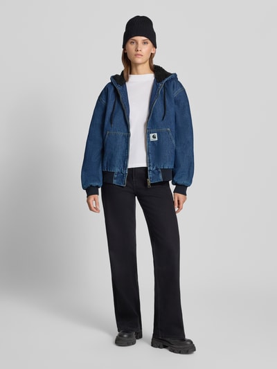 Frauenmodel trägt Jeansjacke mit Kapuze und Reißverschluss von Carhartt Work In Progress, Baumwolle, Blau, Känguru-Tasche, Ganzkörperansicht von vorne.